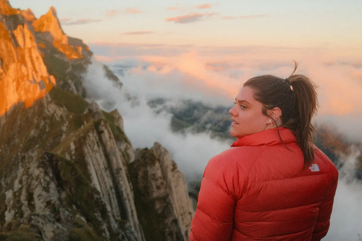 A woman in a red puffer jacket overlooking rocky mountains above the clouds